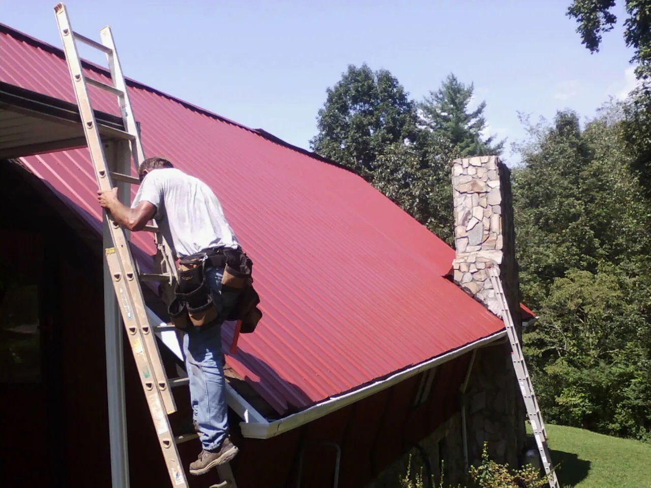 Roofer on a ladder beside a red metal roof and stone chimney — Soffit & Fascia Repair by Roofing Brothers in Weems